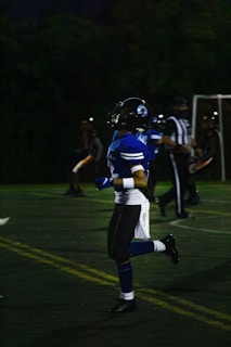 A person in a blue football uniform is running on a field. The player is wearing a helmet with a face guard. Other players and a referee can be seen in the background, all involved in an outdoor sports event during nighttime.