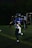 A person in a blue football uniform is running on a field. The player is wearing a helmet with a face guard. Other players and a referee can be seen in the background, all involved in an outdoor sports event during nighttime.