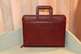 A brown leather briefcase with a rectangular shape and a sturdy handle is positioned upright on a light-colored surface. The background features a decorative patterned curtain in soft beige tones.