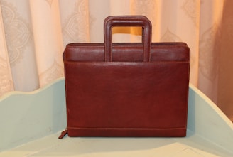 A brown leather briefcase with a rectangular shape and a sturdy handle is positioned upright on a light-colored surface. The background features a decorative patterned curtain in soft beige tones.