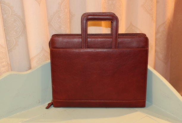 A brown leather briefcase with a rectangular shape and a sturdy handle is positioned upright on a light-colored surface. The background features a decorative patterned curtain in soft beige tones.