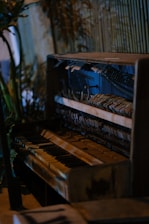 An old, weathered piano with visible wear and tear is situated in a rustic setting. The internal mechanisms of the piano are exposed, showing strings and hammers. The background features wooden slats and some green foliage, adding a natural touch to the scene.
