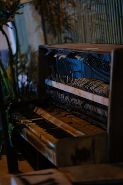 An old, weathered piano with visible wear and tear is situated in a rustic setting. The internal mechanisms of the piano are exposed, showing strings and hammers. The background features wooden slats and some green foliage, adding a natural touch to the scene.