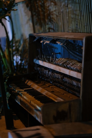 An old, weathered piano with visible wear and tear is situated in a rustic setting. The internal mechanisms of the piano are exposed, showing strings and hammers. The background features wooden slats and some green foliage, adding a natural touch to the scene.