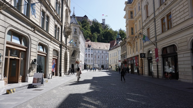 A picturesque street flanked by historic European buildings, with intricate architectural details. The street is paved with cobblestones and features people walking leisurely or riding bicycles. In the background, a castle is visible on a hill, partially obscured by greenery.