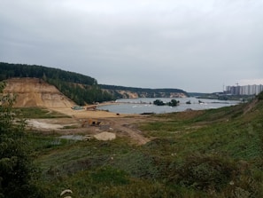 A landscape featuring a construction or mining site near a body of water. The area is surrounded by green vegetation and elevated land. In the background, there are dense trees covering rolling hills. A cluster of tall residential buildings is visible on the right side, and there are several construction vehicles, including an excavator, present on the site.