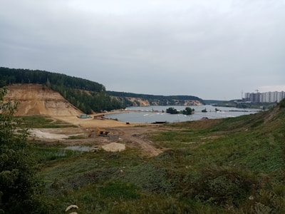 A landscape featuring a construction or mining site near a body of water. The area is surrounded by green vegetation and elevated land. In the background, there are dense trees covering rolling hills. A cluster of tall residential buildings is visible on the right side, and there are several construction vehicles, including an excavator, present on the site.