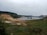 A landscape featuring a construction or mining site near a body of water. The area is surrounded by green vegetation and elevated land. In the background, there are dense trees covering rolling hills. A cluster of tall residential buildings is visible on the right side, and there are several construction vehicles, including an excavator, present on the site.