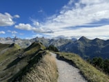 A scenic mountain landscape with a winding hiking trail leading through grassy hills. The trail is surrounded by lush greenery and the scene is set against a backdrop of towering, rugged mountain peaks. The sky is partly cloudy with a bright blue hue.