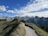 A scenic mountain landscape with a winding hiking trail leading through grassy hills. The trail is surrounded by lush greenery and the scene is set against a backdrop of towering, rugged mountain peaks. The sky is partly cloudy with a bright blue hue.