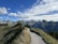 A scenic mountain landscape with a winding hiking trail leading through grassy hills. The trail is surrounded by lush greenery and the scene is set against a backdrop of towering, rugged mountain peaks. The sky is partly cloudy with a bright blue hue.