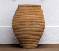 A large ceramic pot with a ribbed texture, featuring a light brown earthy tone. It is positioned against a white textured wall on a wooden floor.