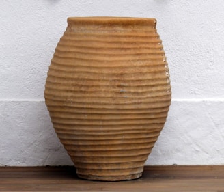 A large ceramic pot with a ribbed texture, featuring a light brown earthy tone. It is positioned against a white textured wall on a wooden floor.