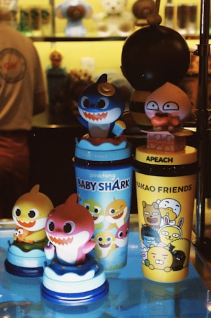 A collection of colorful cartoon-themed cups and figurines displayed on a shelf. Among them are characters from Baby Shark and Kakao Friends, set against a warmly lit backdrop. The figures are vibrant, featuring large, expressive eyes and wide smiles, adding a playful and cheerful vibe.