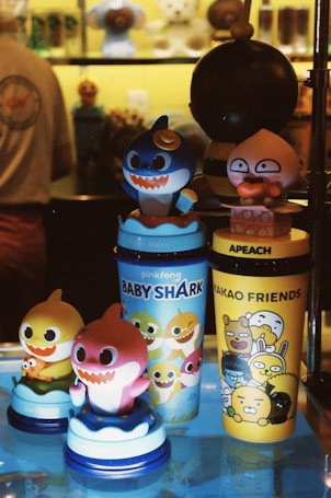 A collection of colorful cartoon-themed cups and figurines displayed on a shelf. Among them are characters from Baby Shark and Kakao Friends, set against a warmly lit backdrop. The figures are vibrant, featuring large, expressive eyes and wide smiles, adding a playful and cheerful vibe.
