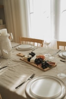A dining table is elegantly set with a cream tablecloth, featuring a wooden serving board displaying an assortment of cheese, black grapes, cured meats, and herbs. Wine glasses and minimalist white plates are arranged neatly alongside cutlery, creating a sophisticated atmosphere.