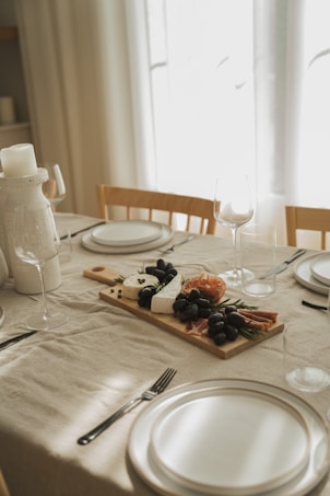 A dining table is elegantly set with a cream tablecloth, featuring a wooden serving board displaying an assortment of cheese, black grapes, cured meats, and herbs. Wine glasses and minimalist white plates are arranged neatly alongside cutlery, creating a sophisticated atmosphere.