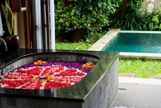 A luxurious spa setting featuring a bathtub filled with vibrant flower petals, arranged in an intricate pattern. The bathtub is situated in an open space with a view of a lush garden and a swimming pool surrounded by greenery. The area exudes a sense of tranquility and relaxation.