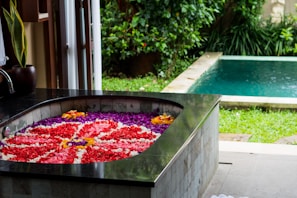 A luxurious spa setting featuring a bathtub filled with vibrant flower petals, arranged in an intricate pattern. The bathtub is situated in an open space with a view of a lush garden and a swimming pool surrounded by greenery. The area exudes a sense of tranquility and relaxation.