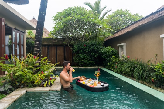 A man is sitting in a private pool surrounded by lush greenery and tropical plants. He is enjoying a drink from a glass while a floating tray with a variety of breakfast items and juices is beside him. The area has a tranquil and luxurious atmosphere with a thatched roof building to the side.