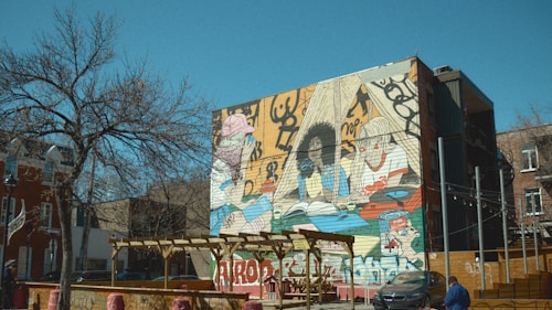 A large mural covers the side of a building, depicting three stylized figures surrounded by various objects and abstract designs. The urban setting includes a bare tree and other brick buildings. In the foreground, there is a wooden pergola with red barrel seating and a car parked on the street.