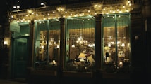 Warm ambient lighting illuminates an inviting cafe interior, visible through large glass windows. The facade features ornate details and vintage decor, with string lights adding a cozy charm. Inside, patrons engage in quiet conversation, surrounded by an eclectic mix of furniture and wall hangings.