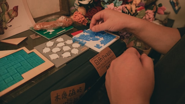 A person arranges puzzle pieces on a small platform surrounded by various types of puzzles and crafts. The setting appears to be a cozy, indoor environment with warm lighting. There are different types of puzzles, including number grids and animal shapes, on a black surface and nearby shelves.