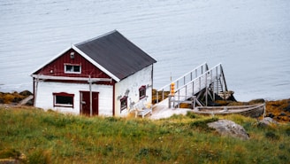 A quaint wooden cabin with a red and white exterior sits on a grassy hill near the water. Attached to the cabin is a wooden walkway that extends over rocks towards the calm water's edge. A modest boat is secured alongside the walkway. The scene is serene and picturesque, suggesting a remote, peaceful location.