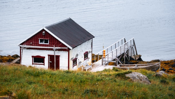 A quaint wooden cabin with a red and white exterior sits on a grassy hill near the water. Attached to the cabin is a wooden walkway that extends over rocks towards the calm water's edge. A modest boat is secured alongside the walkway. The scene is serene and picturesque, suggesting a remote, peaceful location.