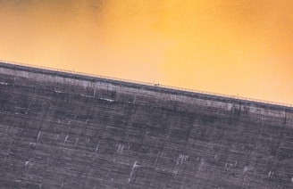 A large dam with a textured concrete surface stretches diagonally across the image. Above the dam, calm water reflects a warm golden hue, likely from the setting or rising sun. Two small figures can be seen walking along the top of the dam, providing a sense of scale to the massive structure.
