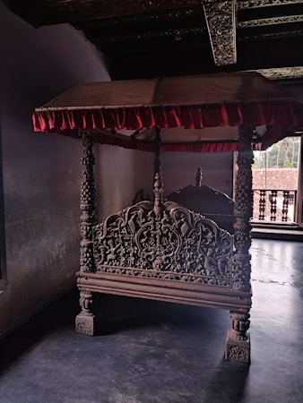 An ornate, intricately carved wooden structure resembling a canopy bed. The bed features detailed carvings on its headboard and posts, and it is adorned with a red fabric canopy. The setting appears historical, with muted lighting and a window showing an outdoor view.