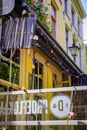 A yellow urban building facade adorned with colorful string lights and adorned with vibrant flowers on the balcony. A glass panel in the foreground displays a logo with a bold letter 'D' and other decorations. Several large windows are visible, as well as a traditional street lamp.