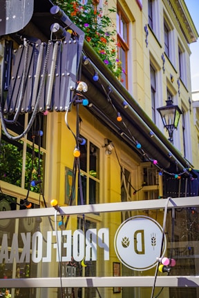 A yellow urban building facade adorned with colorful string lights and adorned with vibrant flowers on the balcony. A glass panel in the foreground displays a logo with a bold letter 'D' and other decorations. Several large windows are visible, as well as a traditional street lamp.