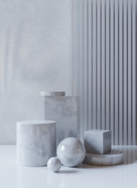 A collection of marble geometric shapes, including cylinders, spheres, and a cube, arranged on a reflective white surface against a textured wall. Vertical stripes add a modern touch.