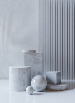 A collection of marble geometric shapes, including cylinders, spheres, and a cube, arranged on a reflective white surface against a textured wall. Vertical stripes add a modern touch.
