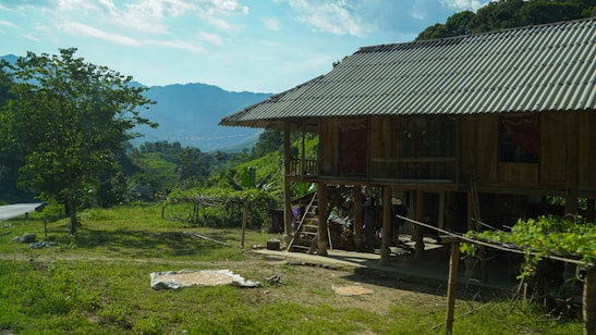 A traditional wooden house raised on stilts in a rural setting, surrounded by lush greenery and mountains. The house features a metal roof and an open area underneath, with some plants and trees nearby. The mountainous background and the clear blue sky contribute to the serene environment.