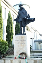 A statue of a man wearing a flowing cape is positioned in front of a modern building with brick walls. The statue is elevated on a pedestal which bears inscriptions including a name and dates. In front of the pedestal, there is a wreath adorned with a bow in the colors of red, white, and green.