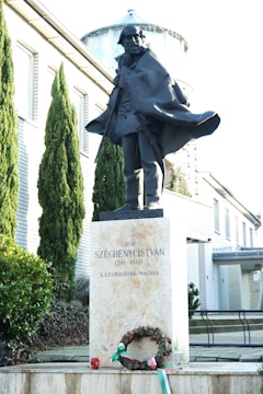 A statue of a man wearing a flowing cape is positioned in front of a modern building with brick walls. The statue is elevated on a pedestal which bears inscriptions including a name and dates. In front of the pedestal, there is a wreath adorned with a bow in the colors of red, white, and green.