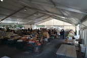 A bustling farmers' market is set up under a large tent with vendors selling a variety of products such as jars of preserves, bottles of sauces, and fresh produce. Wooden tables and racks display the goods, and people are seen browsing and interacting with the sellers. The atmosphere is lively, with natural light filtering through the tent.