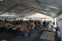 A bustling farmers' market is set up under a large tent with vendors selling a variety of products such as jars of preserves, bottles of sauces, and fresh produce. Wooden tables and racks display the goods, and people are seen browsing and interacting with the sellers. The atmosphere is lively, with natural light filtering through the tent.