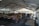 A bustling farmers' market is set up under a large tent with vendors selling a variety of products such as jars of preserves, bottles of sauces, and fresh produce. Wooden tables and racks display the goods, and people are seen browsing and interacting with the sellers. The atmosphere is lively, with natural light filtering through the tent.