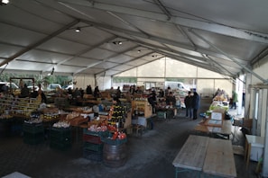 A bustling farmers' market is set up under a large tent with vendors selling a variety of products such as jars of preserves, bottles of sauces, and fresh produce. Wooden tables and racks display the goods, and people are seen browsing and interacting with the sellers. The atmosphere is lively, with natural light filtering through the tent.