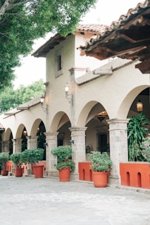 The image features a traditional architectural setting with a row of stone arches supported by columns. Terracotta pots with lush green plants line the walkway, adding a touch of nature. The building features a clay-tiled roof and decorative outdoor lanterns. The ambiance is peaceful and inviting, with trees providing shade in the background.