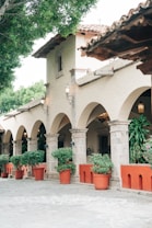 The image features a traditional architectural setting with a row of stone arches supported by columns. Terracotta pots with lush green plants line the walkway, adding a touch of nature. The building features a clay-tiled roof and decorative outdoor lanterns. The ambiance is peaceful and inviting, with trees providing shade in the background.