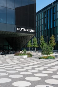 A modern architectural building features a facade with large glass panels and a prominent sign that reads 'Futurium'. In front of the building, there's a plaza with large circular patterns on the ground. Several trees and landscaped areas with greenery and flowers are arranged throughout the plaza. A few people are seated on benches near the building's entrance.