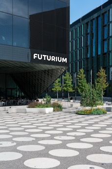 A modern architectural building features a facade with large glass panels and a prominent sign that reads 'Futurium'. In front of the building, there's a plaza with large circular patterns on the ground. Several trees and landscaped areas with greenery and flowers are arranged throughout the plaza. A few people are seated on benches near the building's entrance.