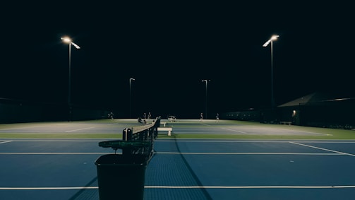 A tennis court is illuminated at night with bright lights. The court surface is blue and green, and there are a few people visible in the distance. A bench and a trash can are positioned in the foreground.