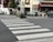 A pedestrian crossing is depicted in an urban area with multiple traffic signs and parked cars. The street is lined with buildings showcasing balconies and decorative flowers. The area appears calm and organized, with a visible cafe or restaurant having people seated inside.