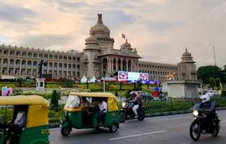 A grand, historic building with a large dome and intricate architecture stands majestically under a cloudy sky. An Indian flag is prominently displayed on top. In the foreground, a busy street features several auto-rickshaws and motorcyclists in motion, suggesting a vibrant city life. Manicured gardens and a statue near the building add to the aesthetic of the scene.