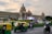 A grand, historic building with a large dome and intricate architecture stands majestically under a cloudy sky. An Indian flag is prominently displayed on top. In the foreground, a busy street features several auto-rickshaws and motorcyclists in motion, suggesting a vibrant city life. Manicured gardens and a statue near the building add to the aesthetic of the scene.